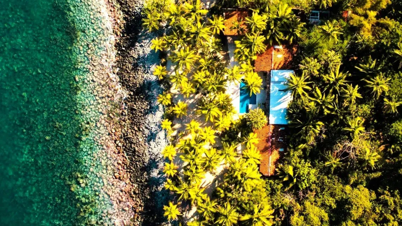 Vista aérea: agua turquesa, orilla rocosa, exuberantes árboles verdes que rodean un edificio con una piscina azul.