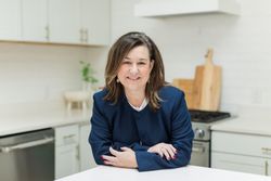 Woman smiling at a kitchen counter in a bright modern kitchen, wearing a navy blazer.