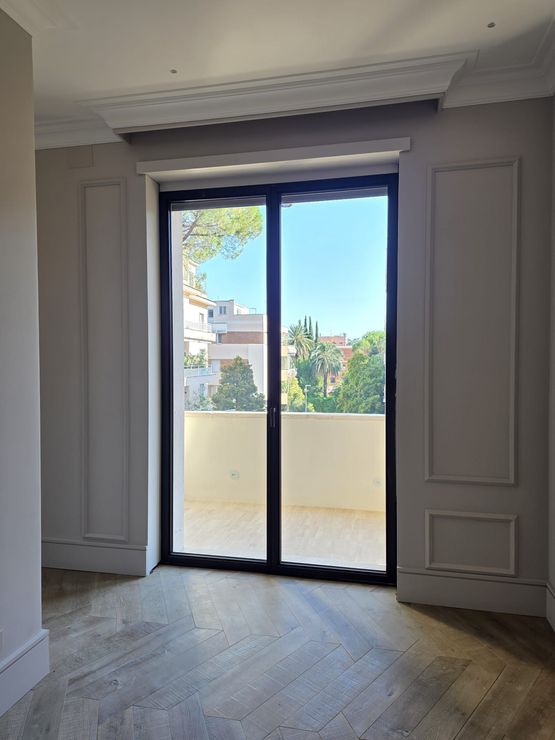 Stanza vuota con pavimento in legno e grandi porte scorrevoli in vetro che si aprono su un balcone e sugli alberi all'esterno.