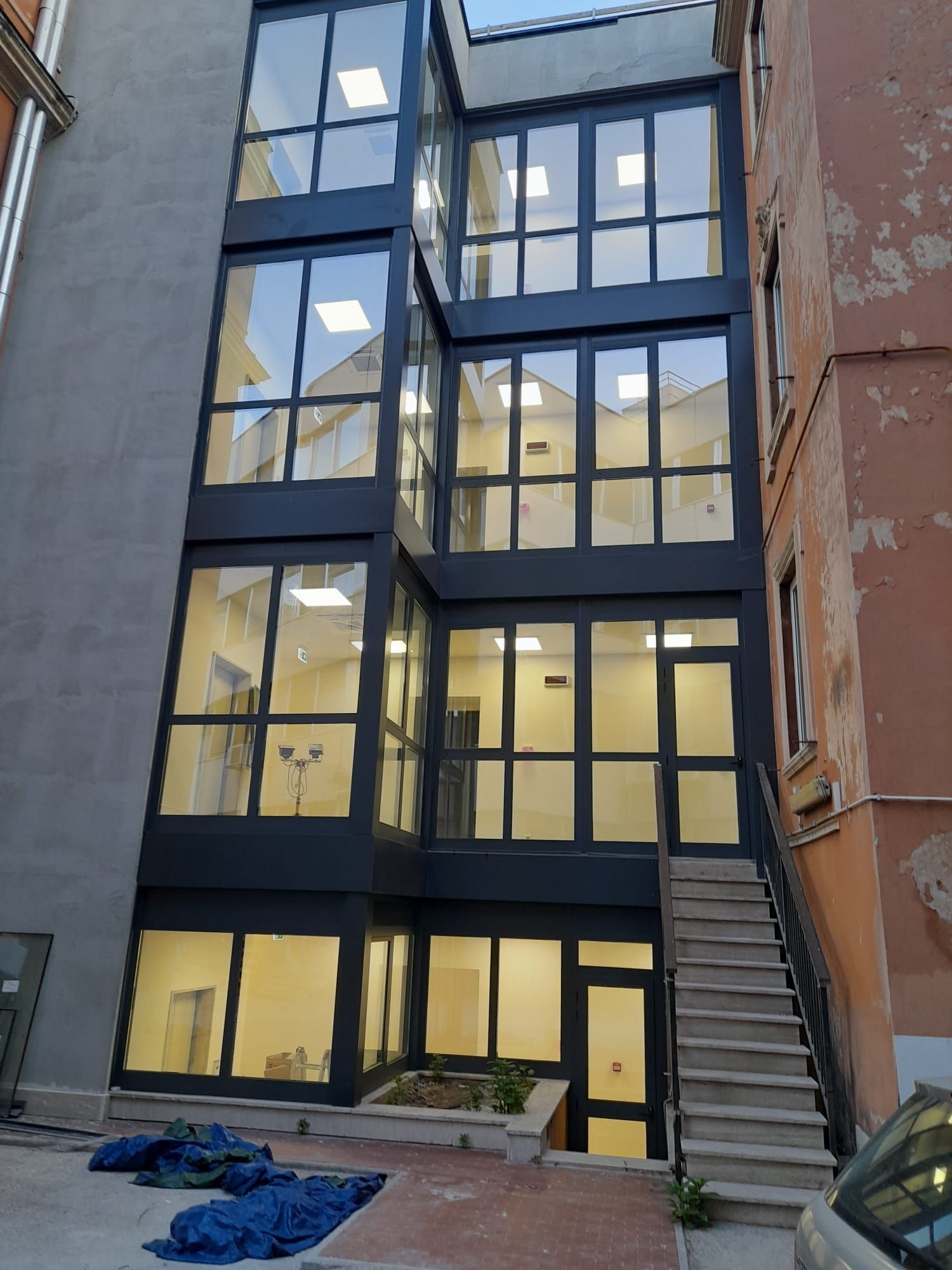 Stretto edificio moderno con facciata in vetro nero, incastonato tra mura più antiche, con scale sulla destra e teloni a terra.