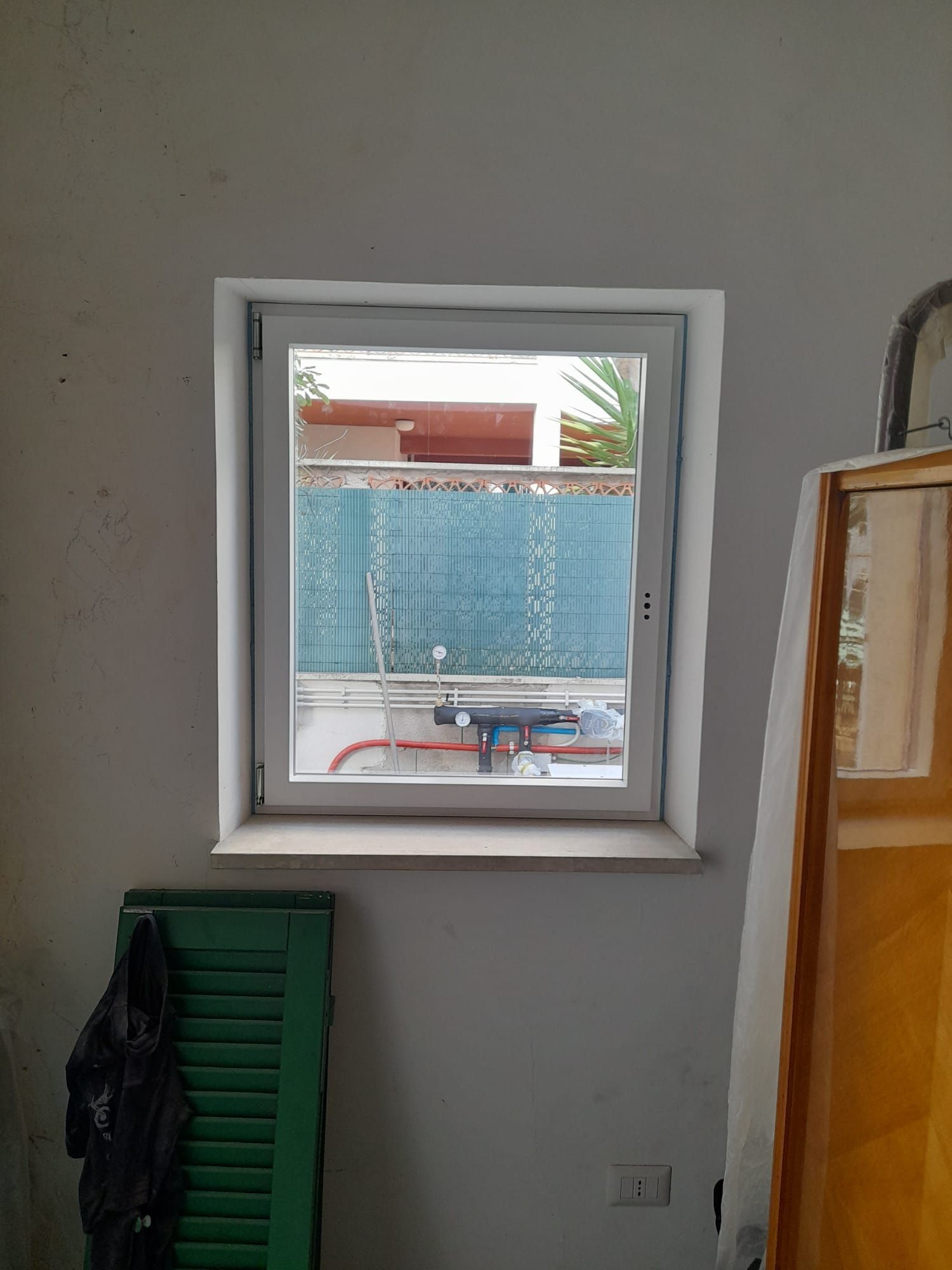 Vista interna dalla finestra su una recinzione blu e un patio, con persiane verdi e un mobiletto in legno nella stanza.