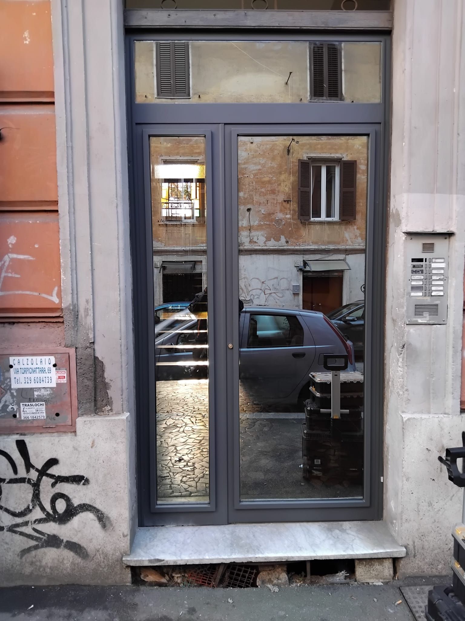 Stretta porta a vetri su un edificio in pietra grigia, che riflette la strada e un'auto parcheggiata.