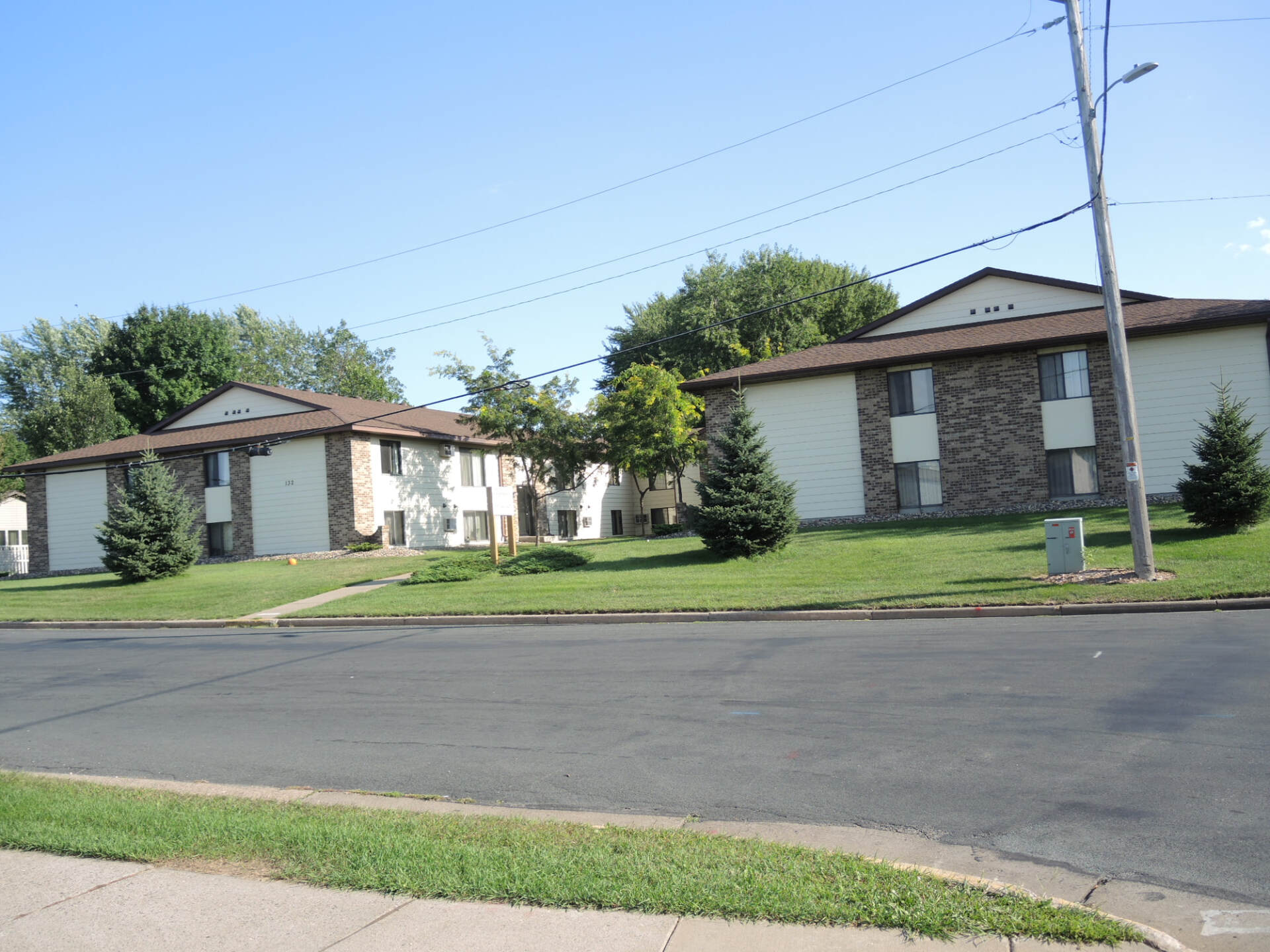 Apartment buildings with beige siding and brown roofs beside a paved street on a sunny day