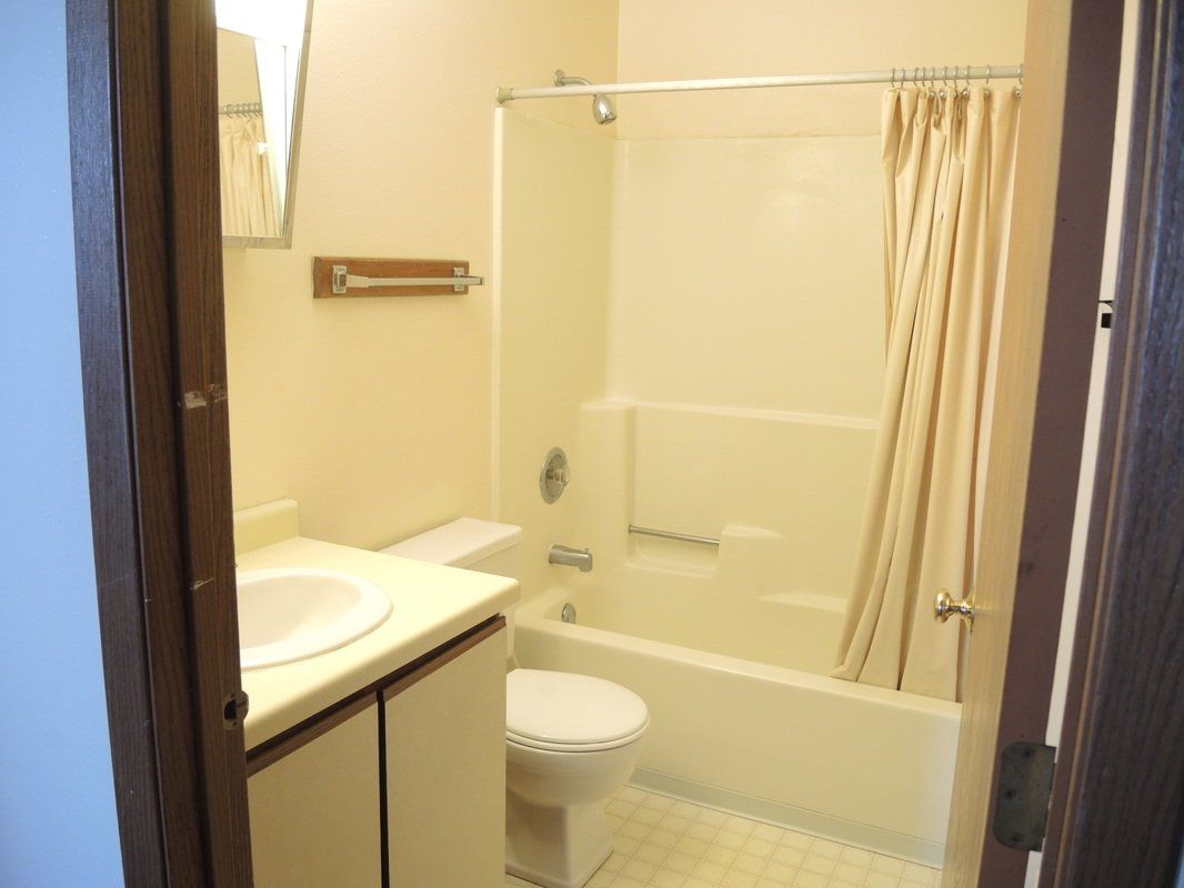 Small beige bathroom with sink, toilet, bathtub, and shower curtain seen through a doorway