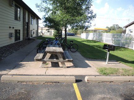 Outdoor picnic table and bike rack beside an apartment building walkway, with a grill and grassy courtyard.