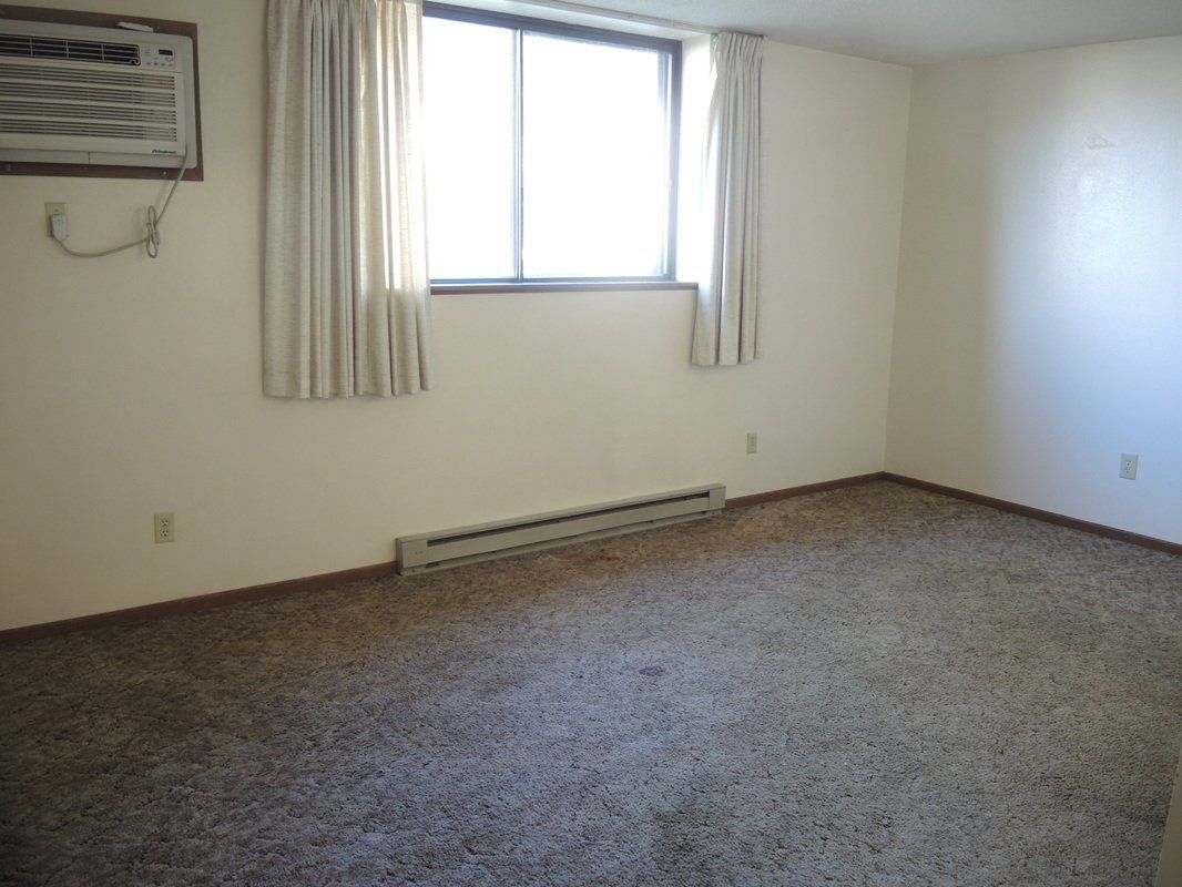 Empty beige room with a window, curtains, wall heater, and brown carpet.