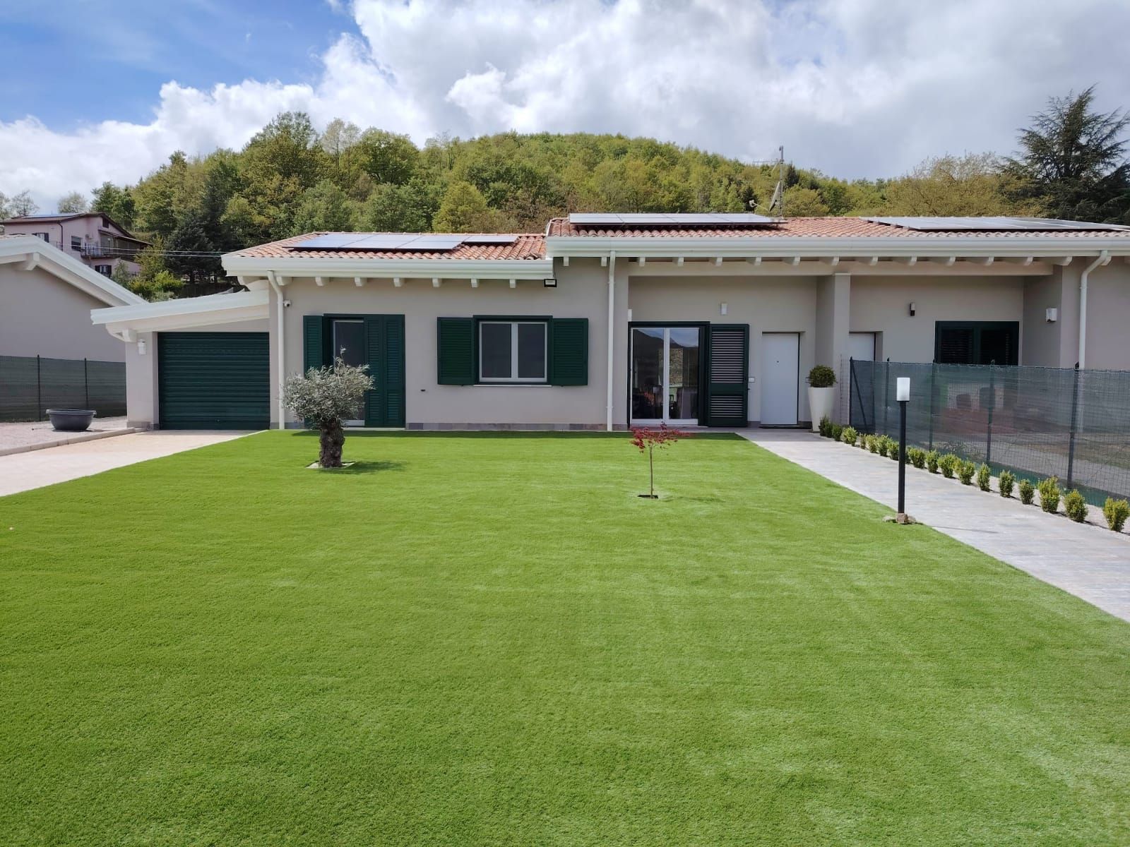 Una casa a un solo piano, di colore chiaro, con persiane verde scuro, un garage, un ampio prato verde e pannelli solari sul tetto.