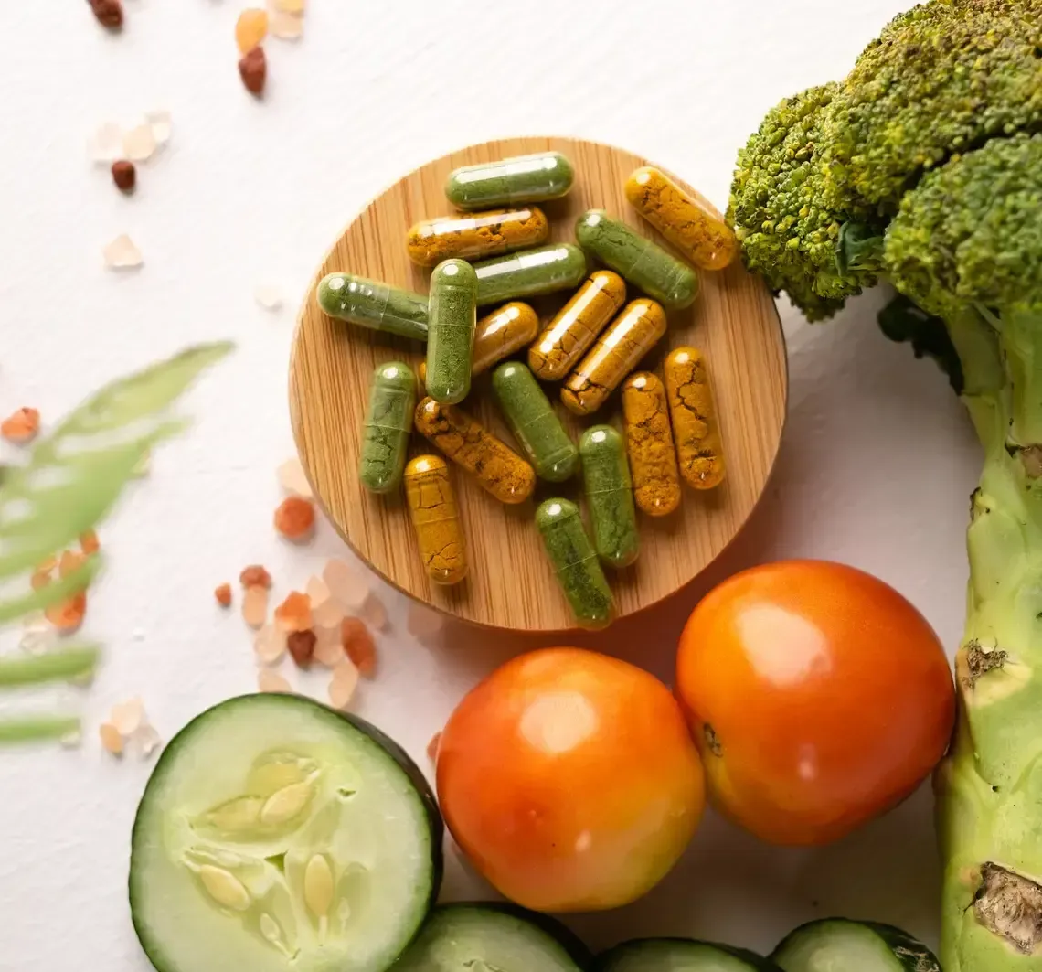 Fresh vegetables and capsules on a white surface, with broccoli, cucumber, and tomatoes surrounding a wooden dish