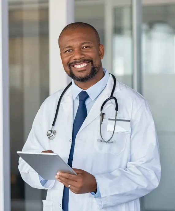 Doctor smiling in white coat with stethoscope, holding a clipboard in a clinic hallway.