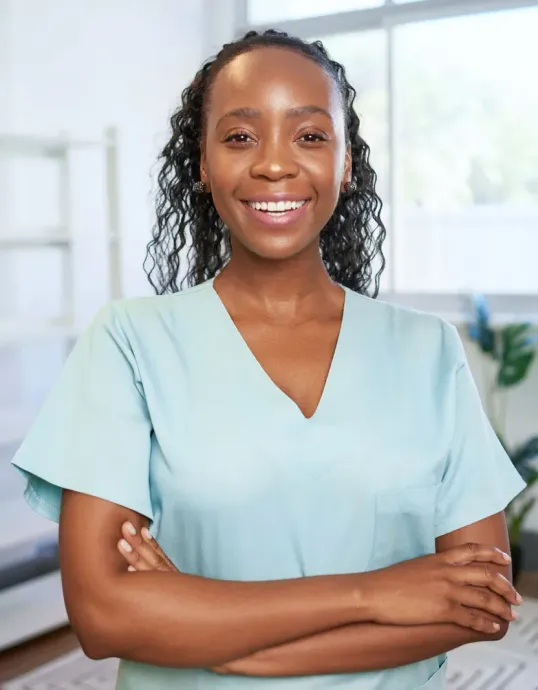 Smiling healthcare professional in a light blue top with arms crossed in a bright office.