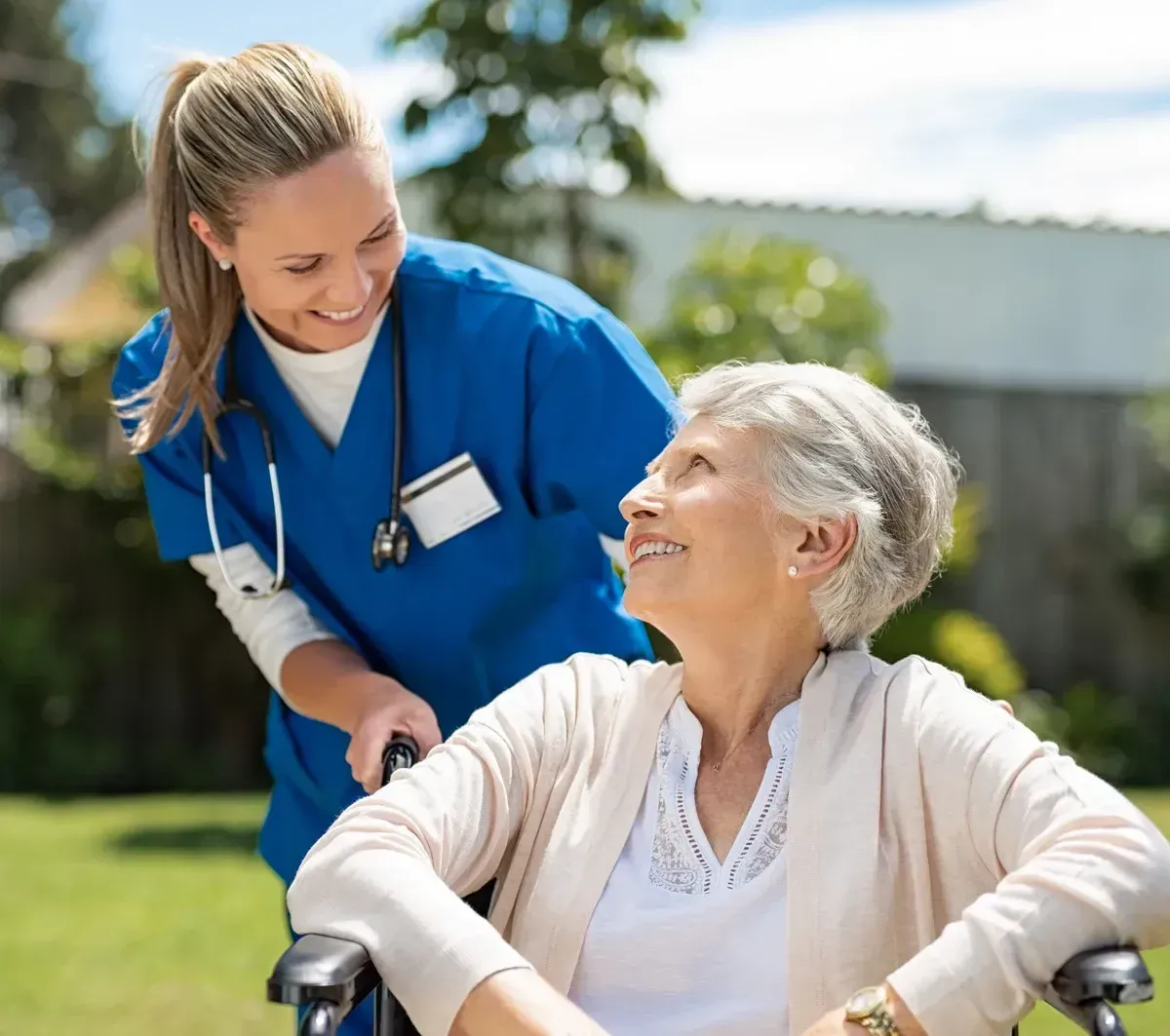 Caregiver in blue scrubs smiles beside a seated patient outdoors in a sunny garden