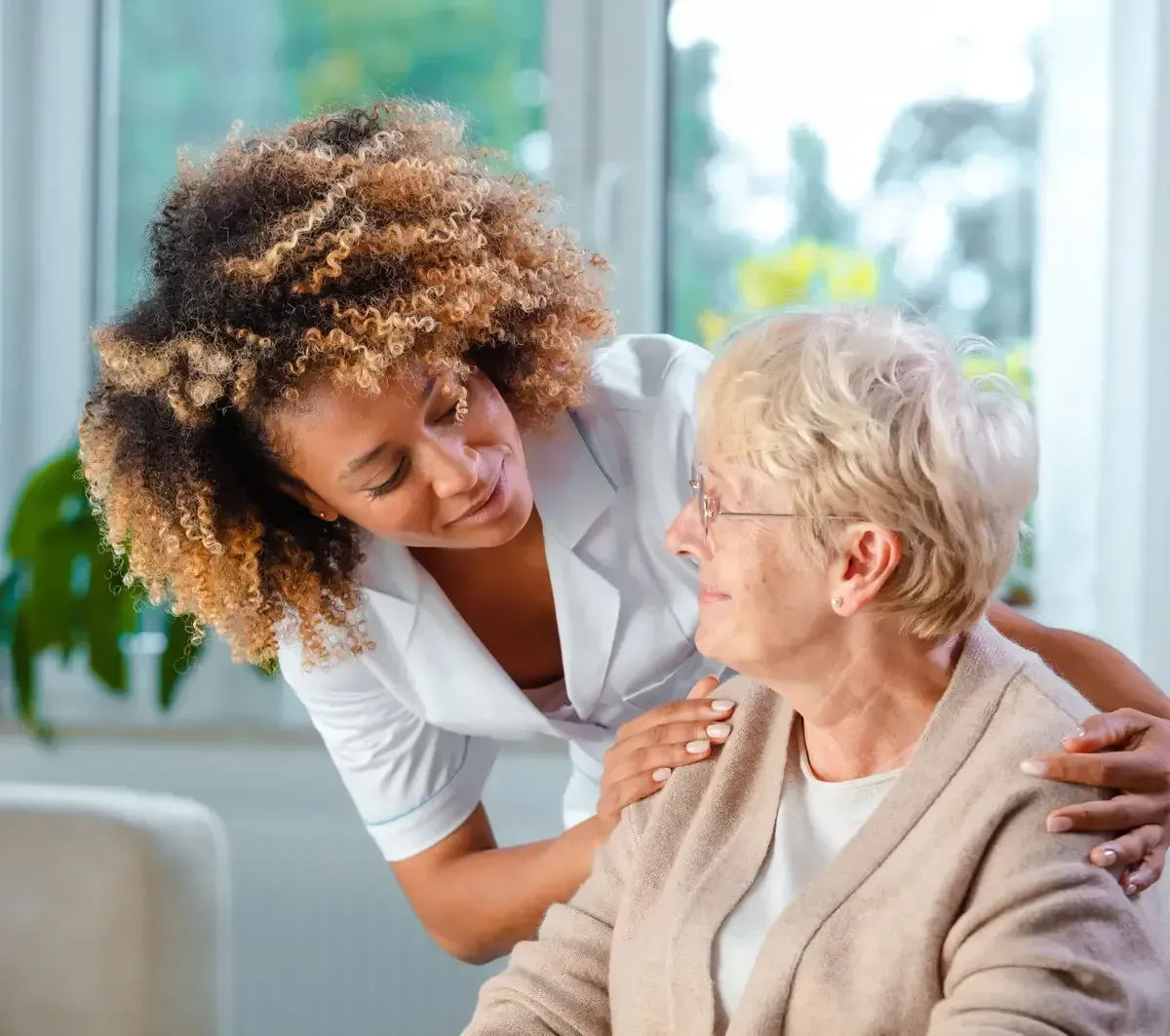 Caregiver gently supporting an older woman indoors by a bright window