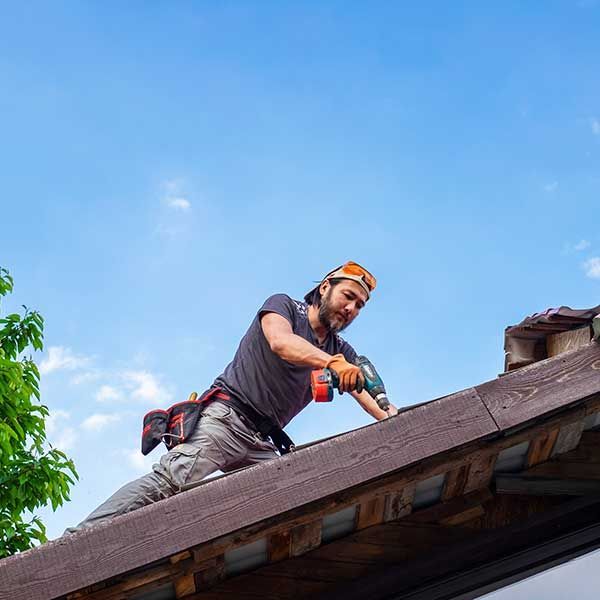 Worker drilling roof shingles on a steep roof under a clear blue sky