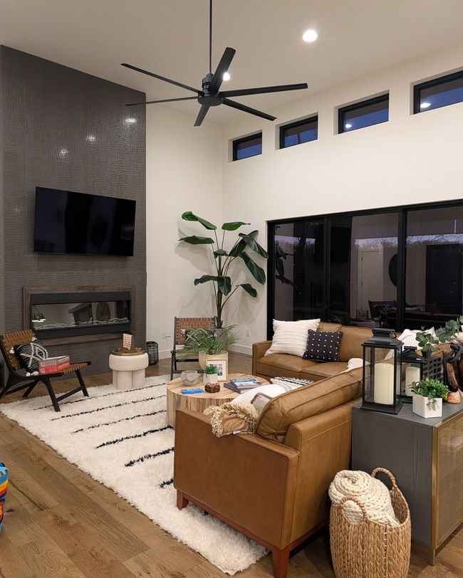 Modern living room with fireplace, wall-mounted TV, tan sofa, large windows, and ceiling fan