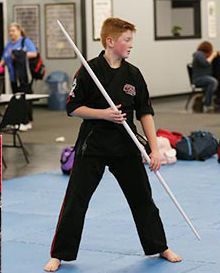 A young boy in a black karate uniform is holding a long pole.