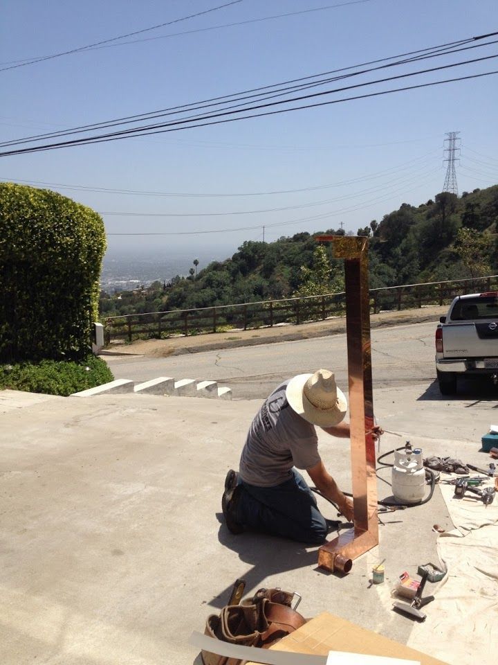 Man in a cowboy hat kneels, working on copper post in driveway. View of trees, hills, and power lines.