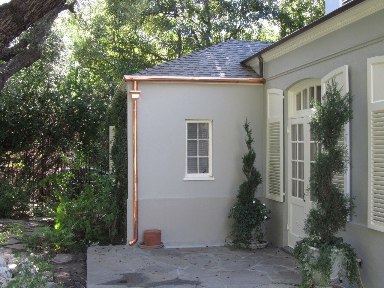 Exterior view of a stucco building with a copper gutter and a small window; trees and greenery surround it.