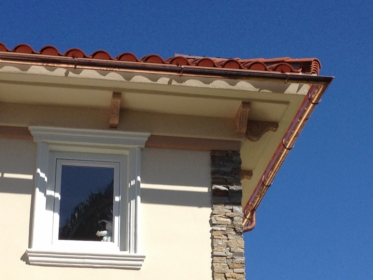 Corner of a stucco house with copper gutters, a tiled roof, window, and stone column against a blue sky.