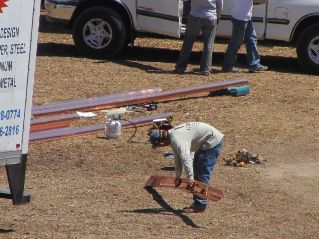 A worker bends over copper sheets on a construction site, with a truck and other workers in the background.