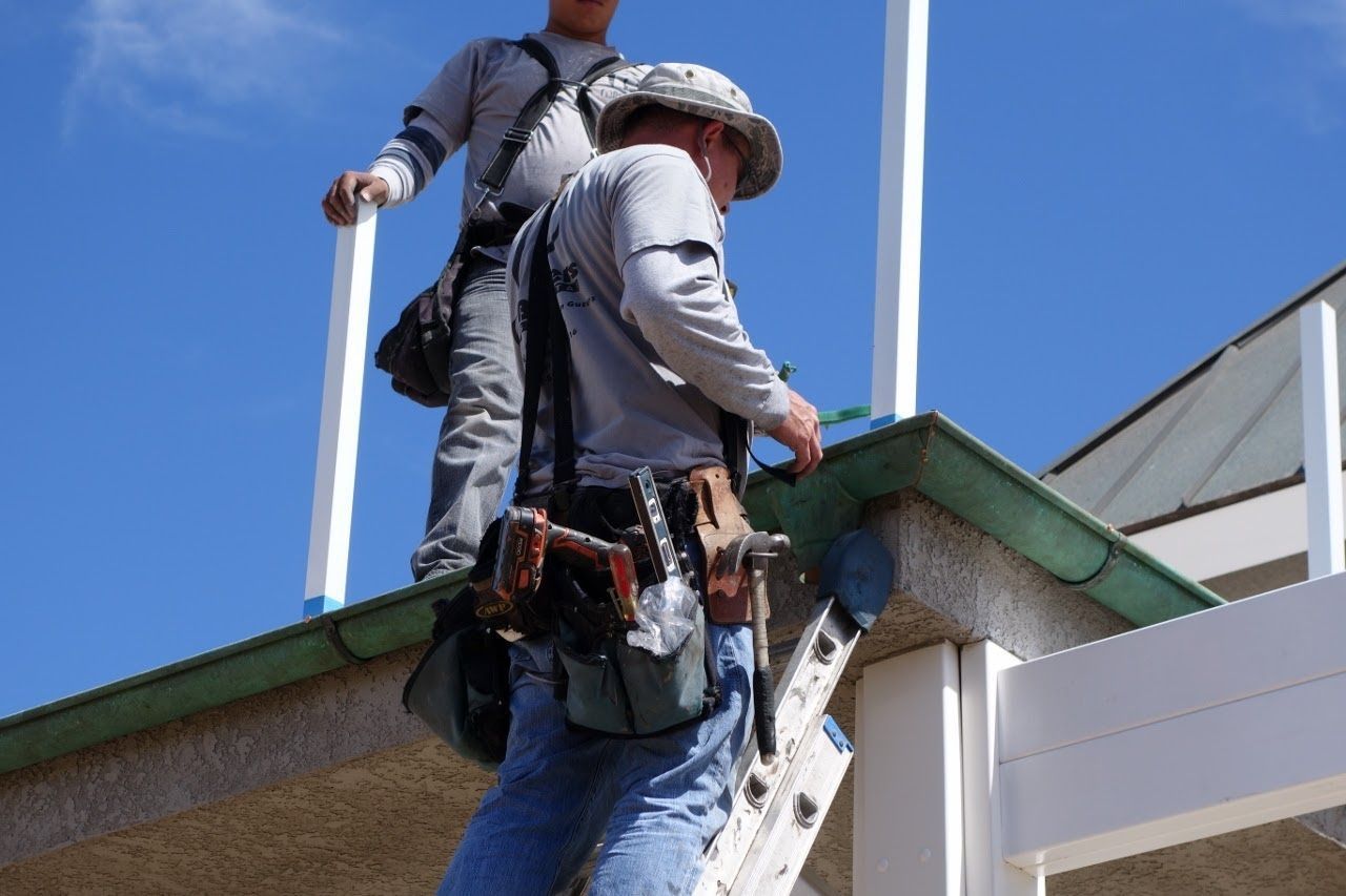 Two construction workers on a roof installing white posts. One wears a tool belt. Blue sky.