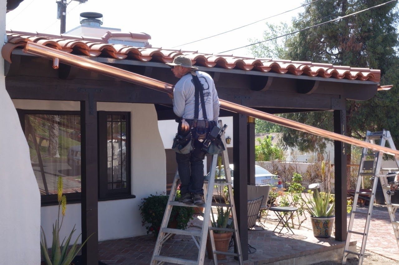 Man on ladder installs gutter on brown patio roof.