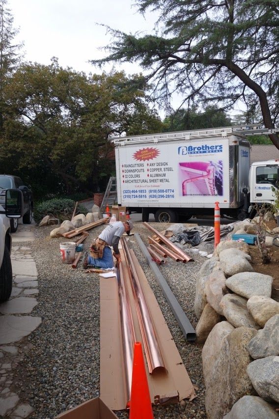 Construction site: Worker with tools near copper pipes, truck, rocks, and gravel driveway.