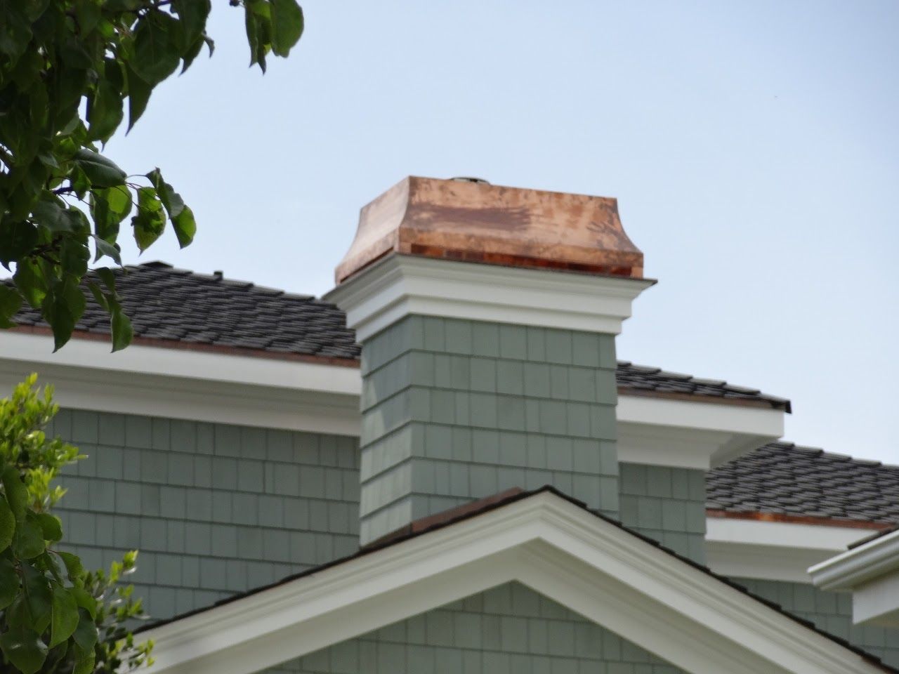 Copper chimney cap on a green shingled chimney, set against a blue sky.