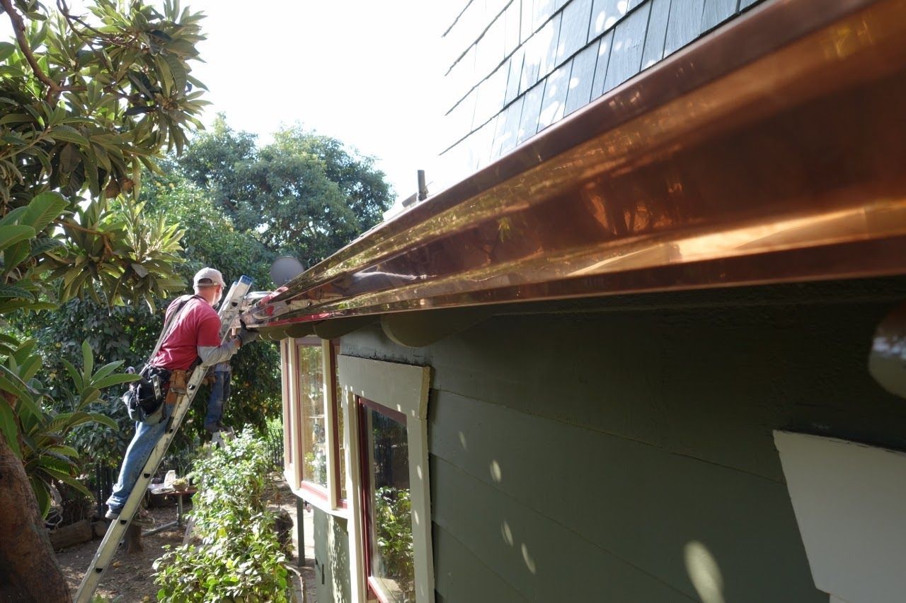 Person on a ladder cleaning copper gutters on a green house with windows; sunny outdoor setting.