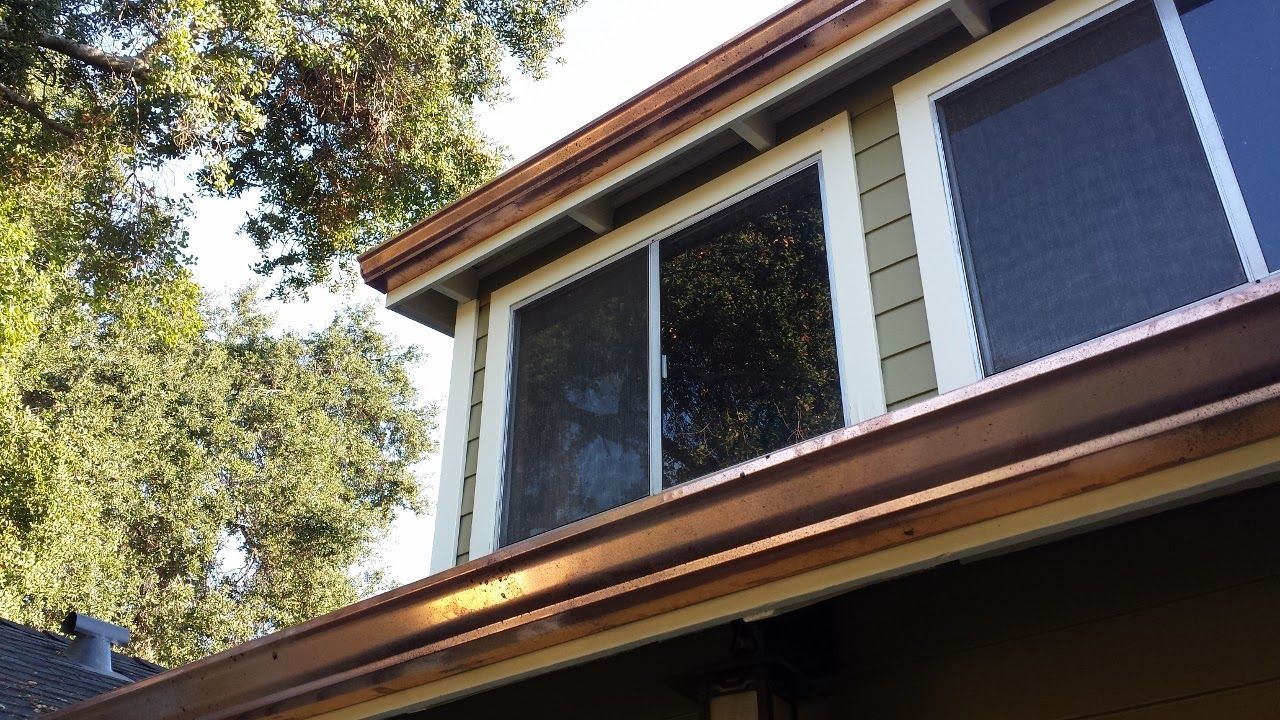 Copper gutters and trim on a house with windows and a backdrop of green trees.