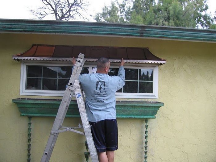 Man on ladder installing a copper awning over a window on a yellow building.