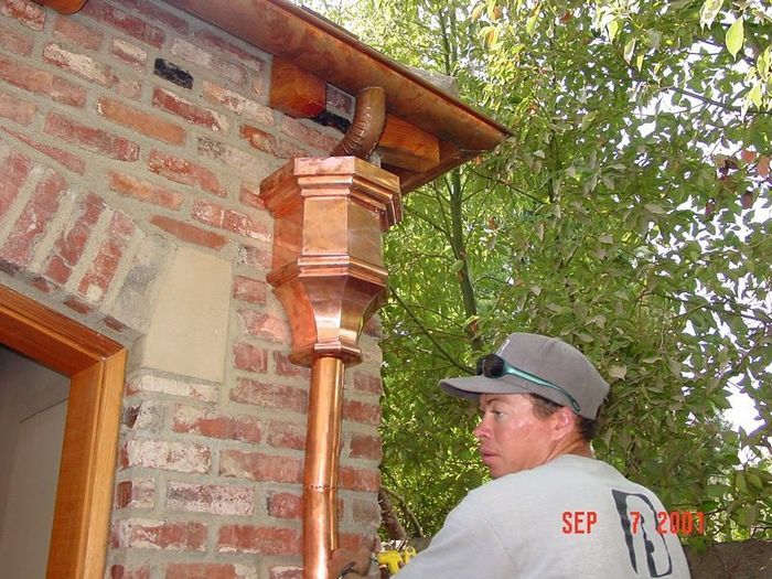 Man in cap, next to a brick building with copper gutter and downspout. Green trees in the background.