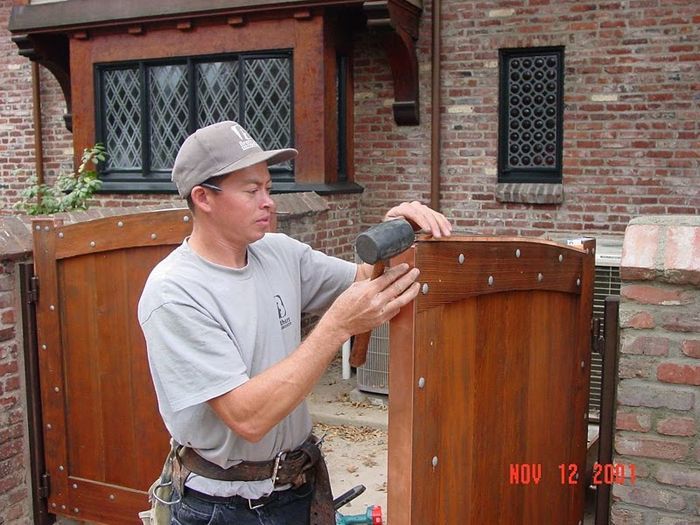 Man installing a wooden gate, using a power tool. Brick building background, autumn.