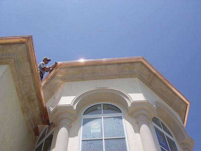Man on roof installing copper trim on a beige building under a blue sky.
