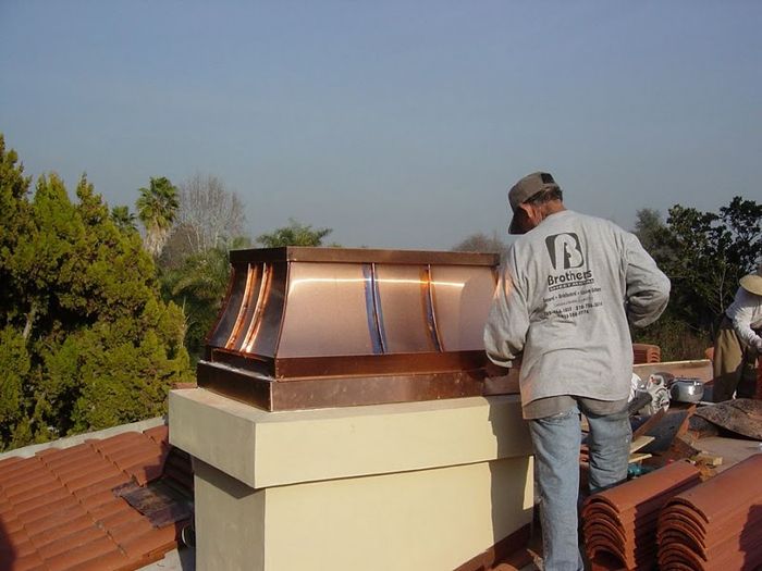 Man installing a copper chimney cap on a rooftop; sunny day, terracotta roof tiles.