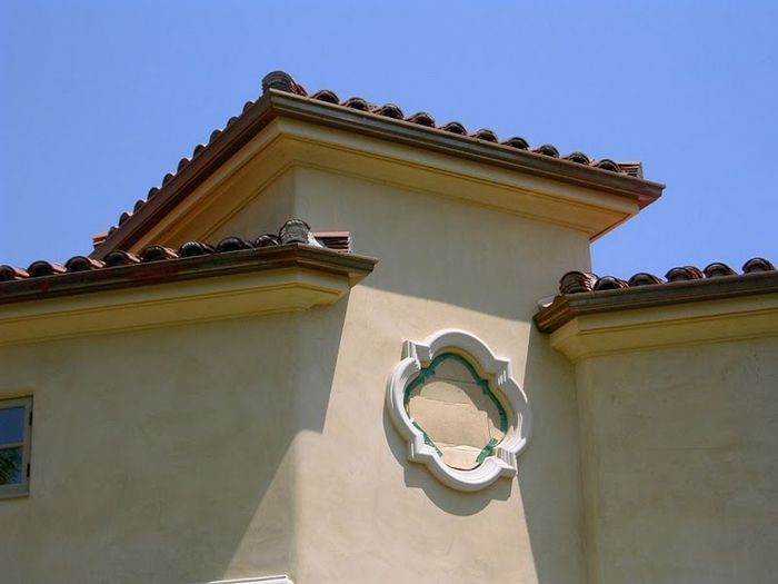 Tan stucco building with red tile roof, decorative window frame, against a blue sky.