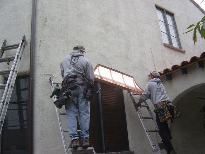 Two workers on ladders installing a copper awning above a doorway on a stucco building.