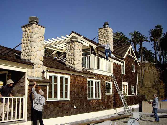 People working on a wood-shingled house with stone chimneys, balcony, and pergola, near a beach with palm trees.