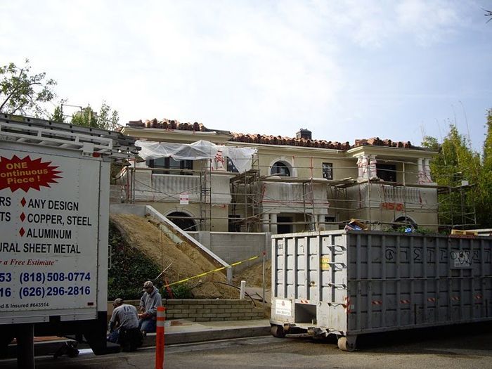 House under construction with scaffolding, workers, and a waste container on a sunny day.