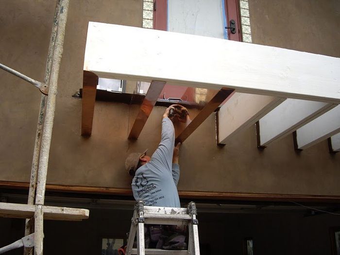 Man on ladder installing wooden pergola beams against a stucco wall.