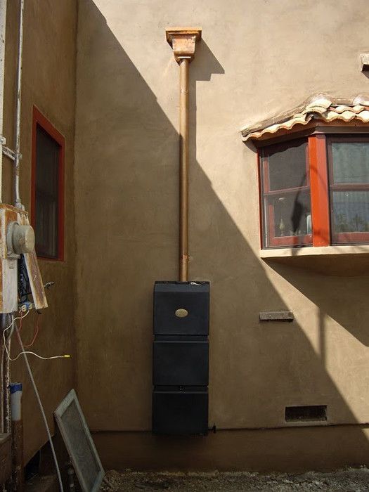 Rainwater harvesting system on a stucco building; copper downspout leads to black storage tanks.
