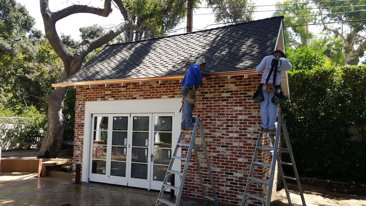 Construction workers on ladders installing roof trim on a brick garage.