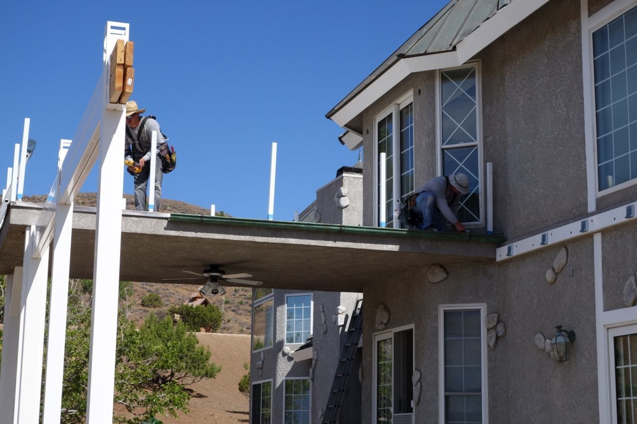 Two workers on a home's roof, installing railings. Beige stucco building, blue sky.