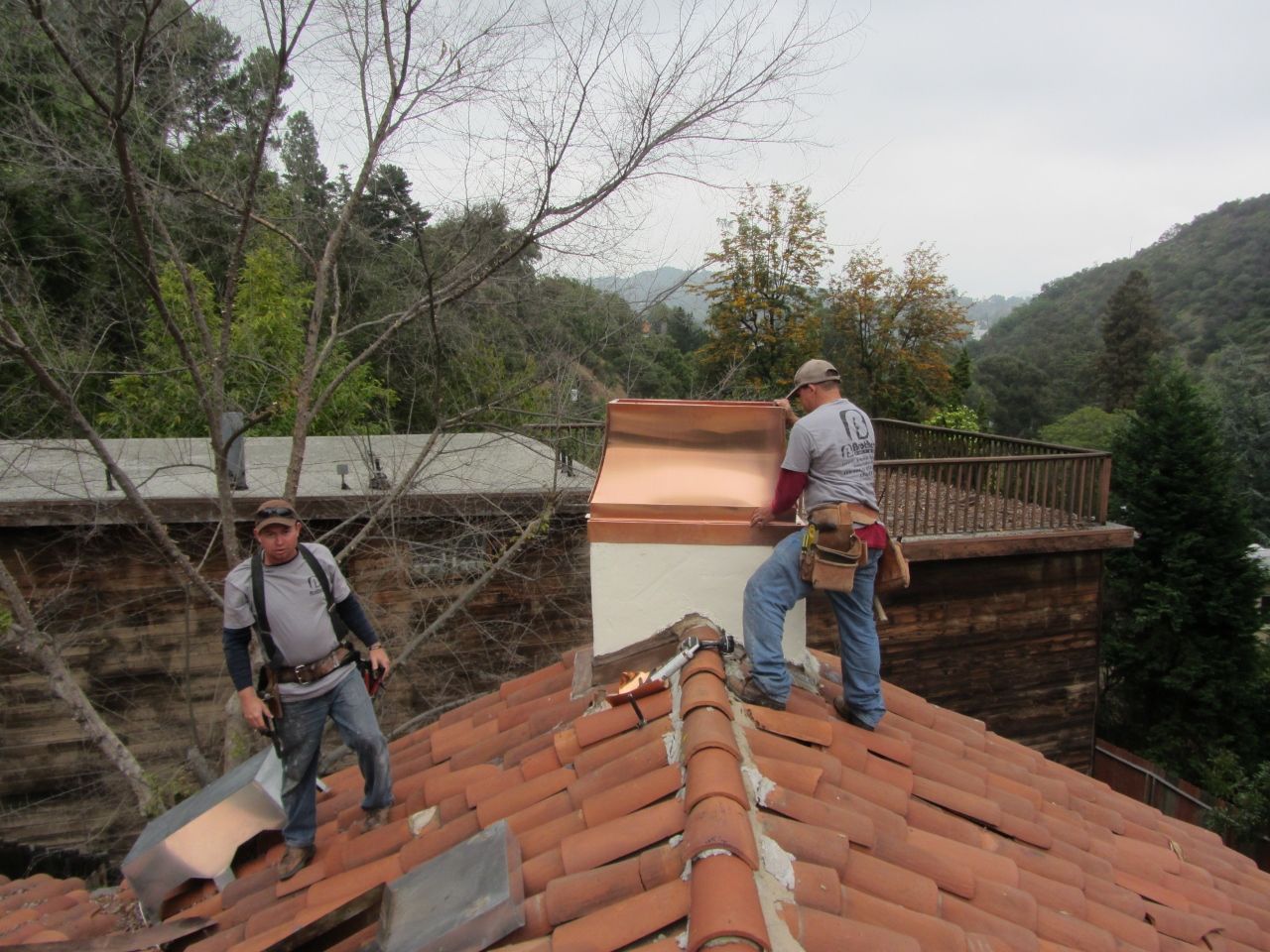 Two roofers on a tiled roof, installing copper flashing around a chimney. Hills in background.