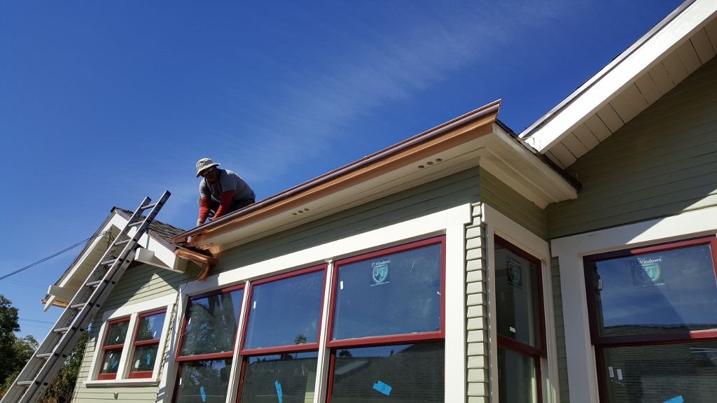 Person on a ladder installing gutters on a house with large windows, under a blue sky.