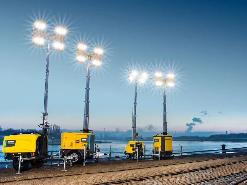 A group of portable lights are sitting on top of a dirt field next to a body of water.