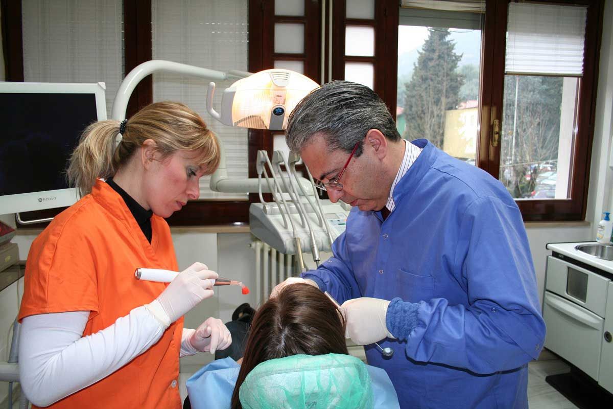 Un dentista e un assistente visitano un paziente in uno studio dentistico, con strumenti e una finestra sullo sfondo.