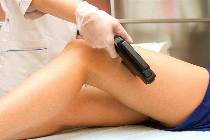 Un operatore sanitario con i guanti utilizza un dispositivo laser portatile sulla gamba di un paziente in un contesto medico.