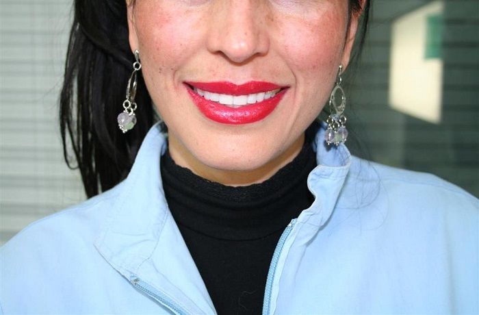 Persona sorridente con rossetto rosso e orecchini d'argento, che indossa una camicia azzurra e un dolcevita nero.
