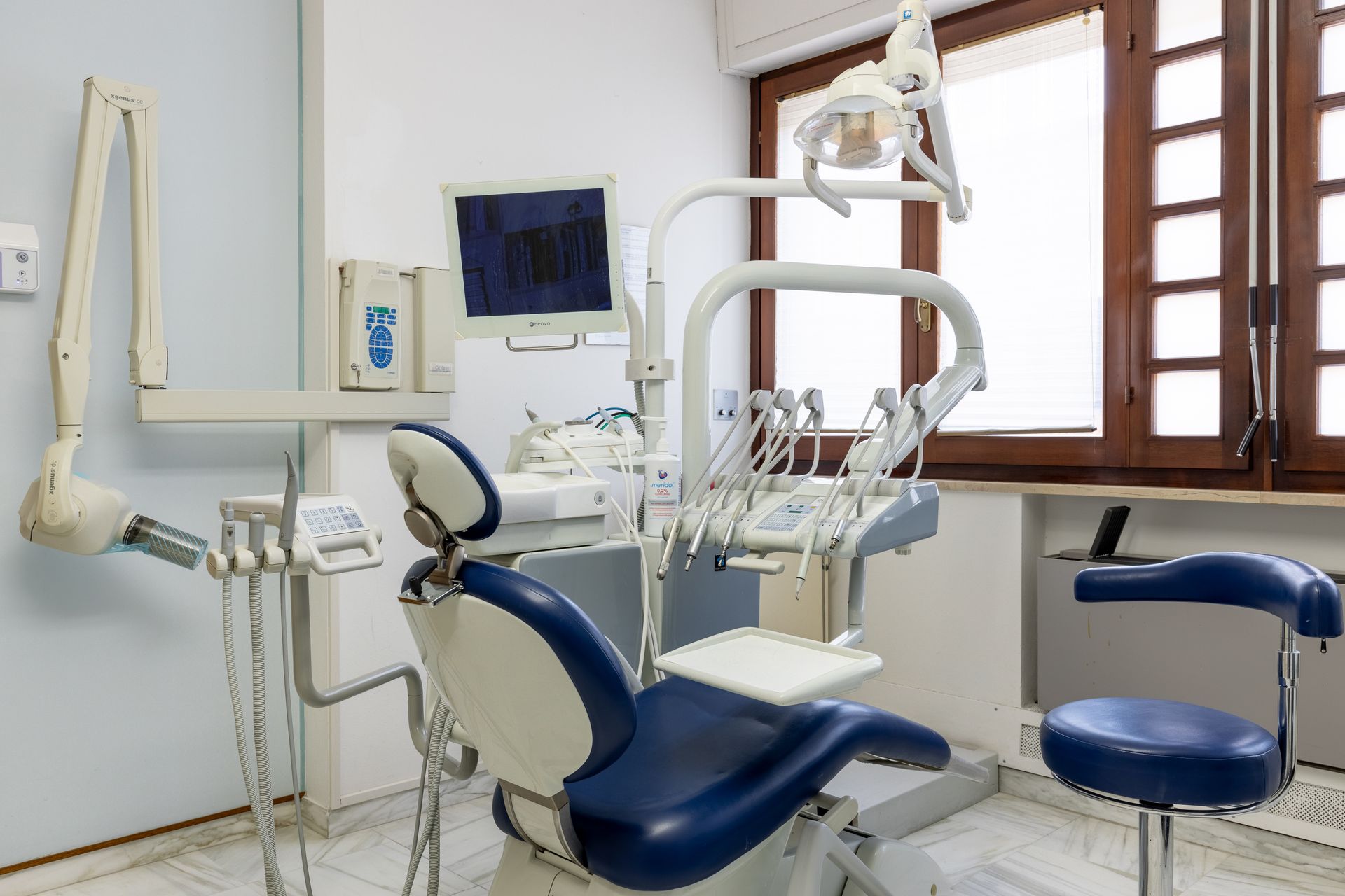 Studio dentistico con poltrona blu per i pazienti, lampade per la visita, monitor e parete vetrata.