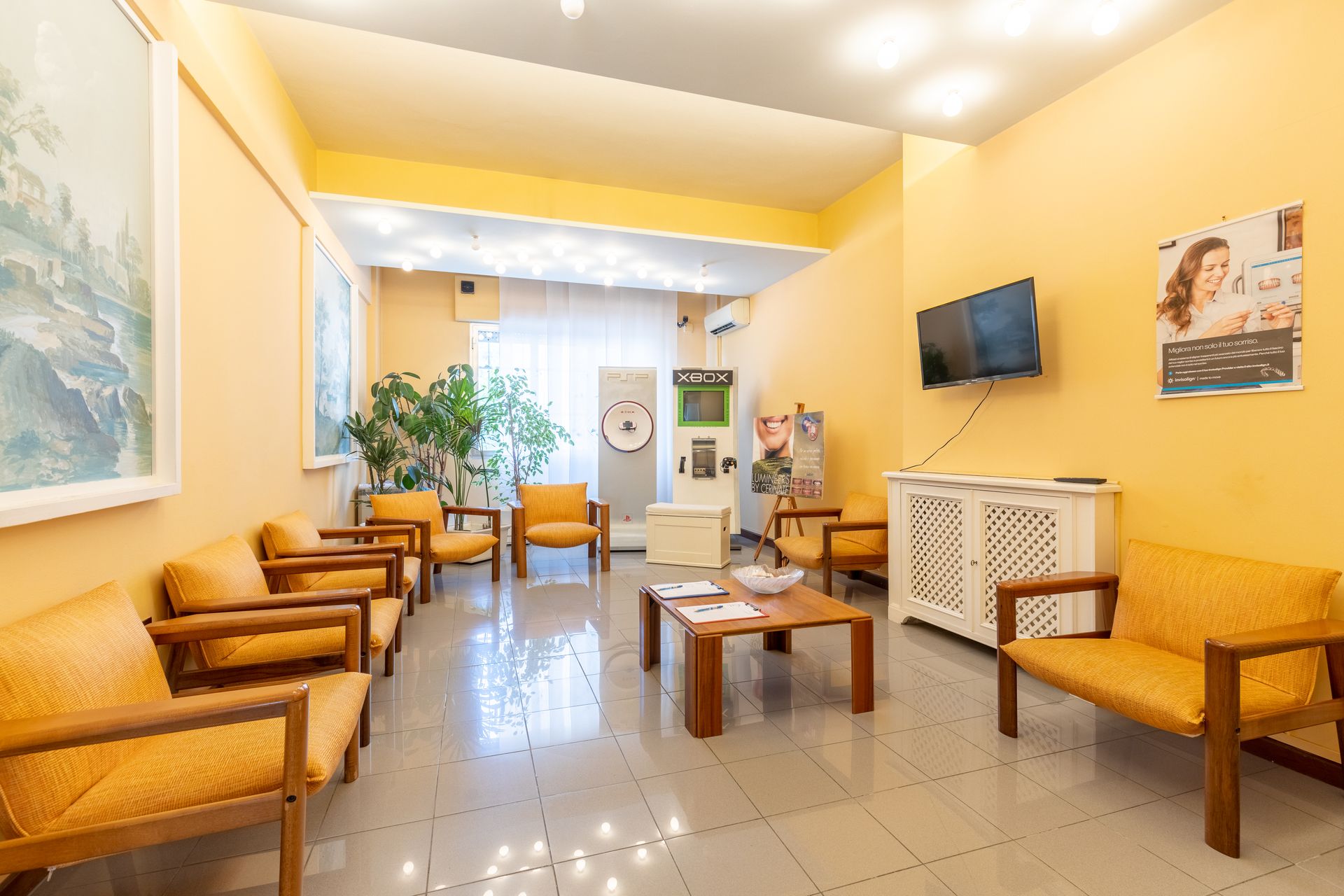 Sala d'attesa luminosa con pareti gialle, sedie in legno, TV, tavolino e pavimento piastrellato.